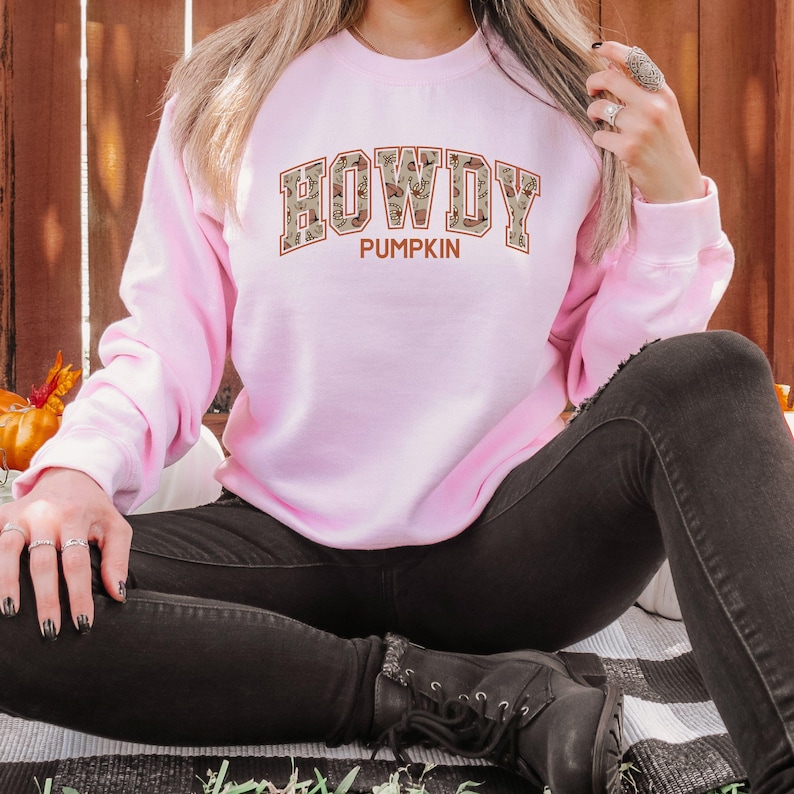 May include: A light pink long-sleeve sweatshirt featuring the text HOWDY PUMPKIN printed on the front. The word HOWDY is displayed in a large, block-style font with a tan and brown patterned fill, while the word PUMPKIN appears below in solid brown.