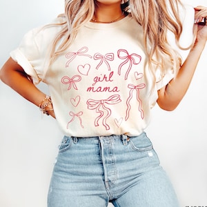 May include: A white t-shirt with a pink graphic design of bows and hearts. The text "girl mama" is in the center of the design.