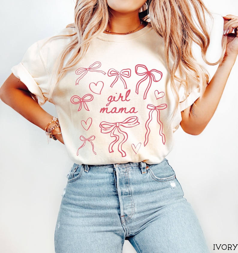 May include: A white t-shirt with a pink graphic design of bows and hearts. The text "girl mama" is in the center of the design.