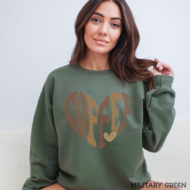May include: Olive green sweatshirt with the word "COFFEE" in a heart shape, using shades of brown and tan. The sweatshirt is a solid color and has long sleeves. The text "MILITARY GREEN" is at the bottom.