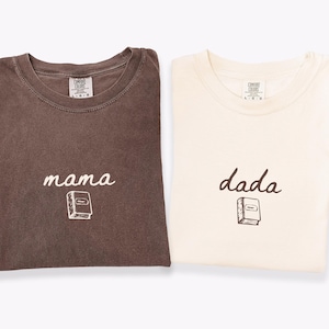 May include: Two crewneck t-shirts, one in espresso brown and one in ivory. The espresso shirt features the word mama in white script above a small book graphic. The ivory shirt features the word dada in brown script above a small book graphic.
