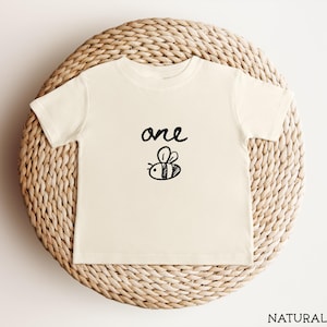 May include: A cream-coloured toddler t-shirt featuring the word "one" above a black cartoon bee illustration. The shirt is made of soft material and is displayed on a woven, circular surface.