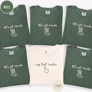 May include: Five short-sleeved crewneck t-shirts. Four shirts are moss green with white embroidered text reading let's get rowdy and a cowboy boot graphic. One shirt is ivory with black embroidered text reading my last rodeo and a diamond ring graphic.