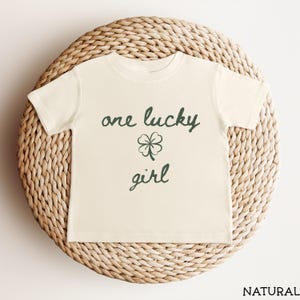 May include: A cream-colored t-shirt with the text "one lucky girl" in dark green script, along with a four-leaf clover graphic. The shirt is displayed on a woven, circular surface.