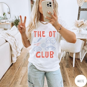 Peut inclure: Un t-shirt blanc &agrave; manches courtes avec le texte rouge THE OT CLUB. Sous le texte se trouvent des graphismes au trait bleu incluant un crayon, deux pi&egrave;ces de puzzle, un soleil souriant, un cerveau et un arc-en-ciel avec le texte rouge every milestone matters.
