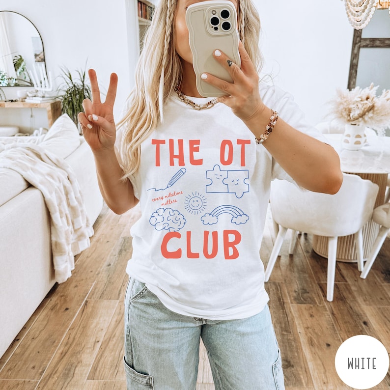 Peut inclure: Un t-shirt blanc &agrave; manches courtes avec le texte rouge THE OT CLUB. Sous le texte se trouvent des graphismes au trait bleu incluant un crayon, deux pi&egrave;ces de puzzle, un soleil souriant, un cerveau et un arc-en-ciel avec le texte rouge every milestone matters.