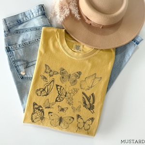 May include: A mustard yellow t-shirt with a graphic print of black butterflies. The shirt has a crew neck and short sleeves. The text "COMFORT COLORS" and "XS-TO-3XL" is printed on the tag.