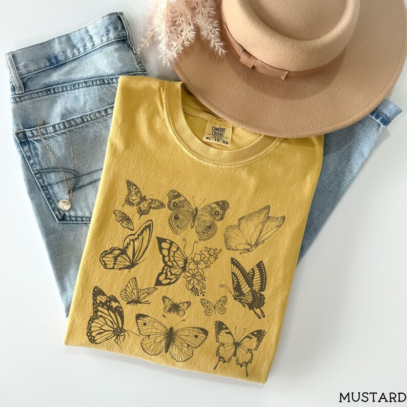 May include: A mustard yellow t-shirt with a graphic print of black butterflies. The shirt has a crew neck and short sleeves. The text "COMFORT COLORS" and "XS-TO-3XL" is printed on the tag.
