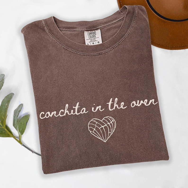 May include: A brown Comfort Colors t-shirt with the phrase "conchita in the oven" in white script, along with a white heart-shaped concha graphic. A brown hat is visible in the background.
