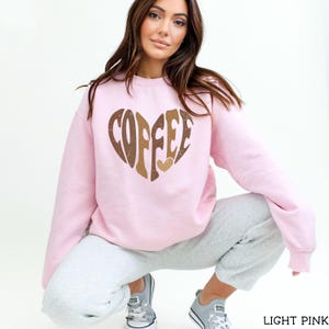 May include: Light pink sweatshirt with a heart-shaped graphic that says "COFFEE" in brown and gold lettering. The sweatshirt is a crew neck style and is made of a soft material. The model is wearing gray sweatpants and gray sneakers.