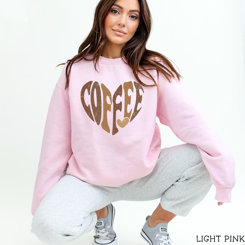May include: Light pink sweatshirt with a heart-shaped graphic that says "COFFEE" in brown and gold lettering. The sweatshirt is a crew neck style and is made of a soft material. The model is wearing gray sweatpants and gray sneakers.