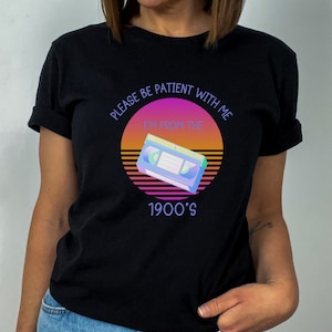 May include: A black t-shirt featuring a graphic of a VHS tape centered within a sunset-colored circle. The text reads, "PLEASE BE PATIENT WITH ME I'M FROM THE 1900'S" in a light purple font. The design incorporates horizontal lines and a retro color palette.