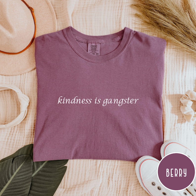 May include: A berry-colored t-shirt with the phrase "kindness is gangster" printed in white. The shirt is a crew neck style and is displayed on a cream-colored surface. White sneakers with red accents are visible.