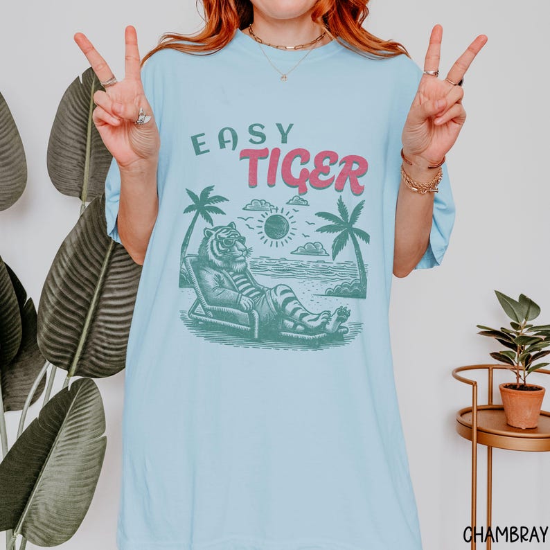 May include: A light blue t-shirt with the text "Easy Tiger" in pink and green. The graphic features a cartoon tiger relaxing on a beach chair with palm trees and a sun in the background.