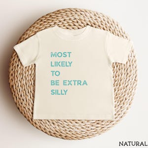 Puede incluir: Una camiseta de manga corta de color crema con el texto "MOST LIKELY TO BE EXTRA SILLY" impreso en una fuente azul claro desgastada. La prenda cuenta con un cuello redondo y un corte est&aacute;ndar, mostrando un dise&ntilde;o sencillo y limpio.