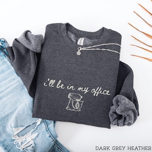 May include: A dark grey heather crewneck sweatshirt featuring the white text "i'll be in my office" above a line art illustration of a stand mixer. The garment has long sleeves with ribbed cuffs and a Gildan brand label at the inner neckline.