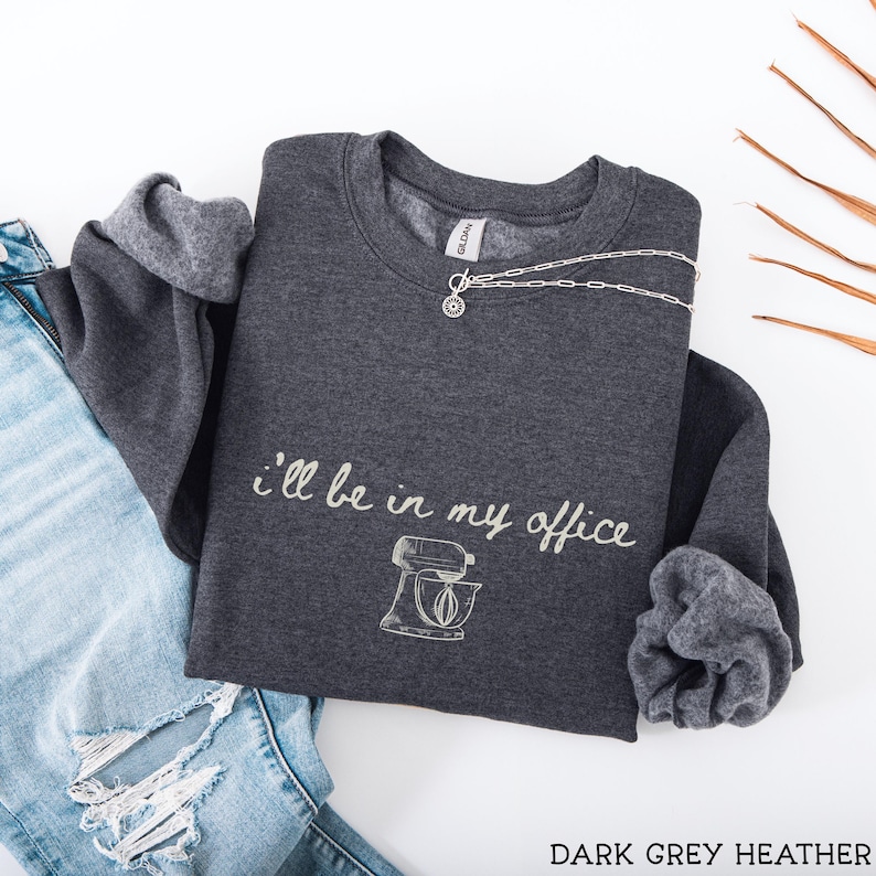 May include: A dark grey heather crewneck sweatshirt featuring the white text "i'll be in my office" above a line art illustration of a stand mixer. The garment has long sleeves with ribbed cuffs and a Gildan brand label at the inner neckline.