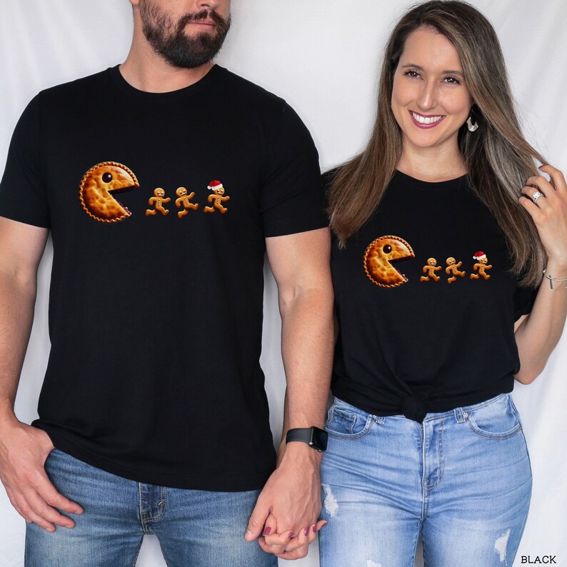 Pu&ograve; includere: Una t-shirt nera a maniche corte con una stampa grafica raffigurante una torta dorata a forma di personaggio di un videogioco che insegue tre piccoli biscotti di pan di zenzero, uno dei quali indossa un cappellino natalizio rosso e bianco.