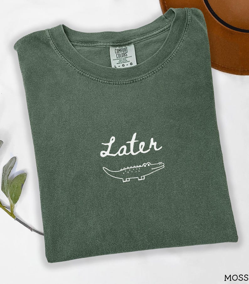 Pu&ograve; includere: Una maglietta verde muschio piegata con la scritta "Later" in bianco sopra un'illustrazione di un alligatore bianco. La maglietta ha un'etichetta Comfort Colors.