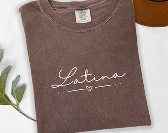 May include: A brown t-shirt with the text "Latina" in white cursive lettering with a small heart and a line underneath. The shirt is made of a soft, comfortable material and is perfect for everyday wear.