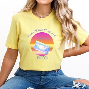 May include: A yellow short-sleeve t-shirt featuring a graphic of a VHS cassette tape centered over a sunset-inspired circle. The text reads, "PLEASE BE PATIENT WITH ME I'M FROM THE 1900'S" in blue lettering. The shirt is paired with blue denim jeans.