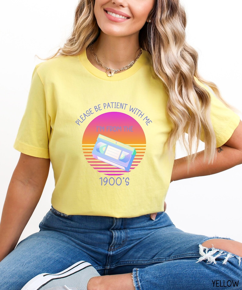 May include: A yellow short-sleeve t-shirt featuring a graphic of a VHS cassette tape centered over a sunset-inspired circle. The text reads, "PLEASE BE PATIENT WITH ME I'M FROM THE 1900'S" in blue lettering. The shirt is paired with blue denim jeans.