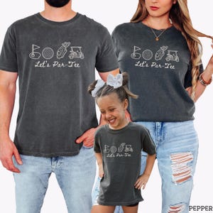 May include: Three dark gray t-shirts with the text "Let's Par-Tee" and golf-themed graphics. The shirts are worn by three people, with the text and graphics in white. The shirts are made of a soft, comfortable material.