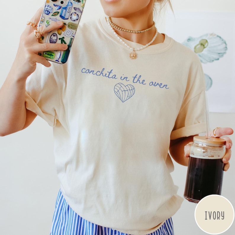 May include: Ivory-colored t-shirt with the text "conchita in the oven" in blue script, along with a blue heart graphic. The shirt is a relaxed fit. The person is holding a phone and a glass of dark liquid.