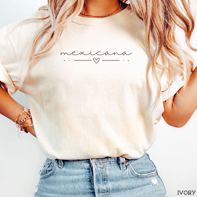 May include: A white t-shirt with the text "mexicana" in a cursive font with a small heart between two lines. The shirt is being worn by a person with long blonde hair and blue jeans.