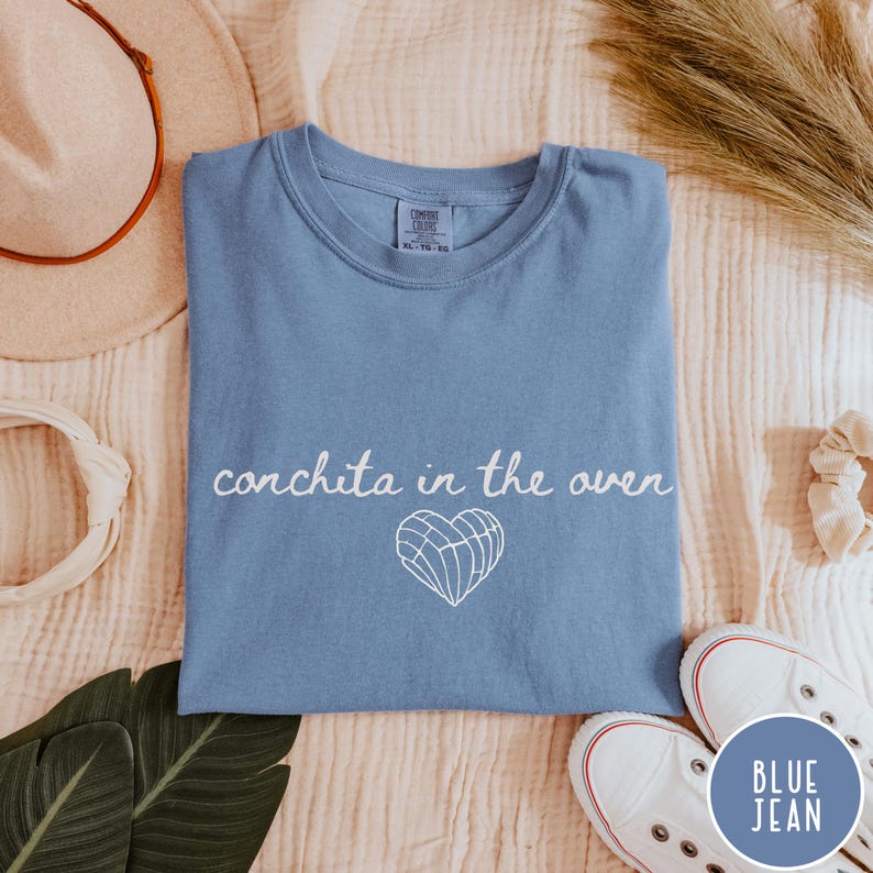May include: A blue jean colored t-shirt with the phrase "conchita in the oven" in white script. A white heart-shaped graphic is below the text. The shirt is laid flat with a pair of white sneakers and a beige hat.