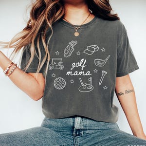 Puede incluir: Una camiseta gris con un dise&ntilde;o gr&aacute;fico blanco de art&iacute;culos relacionados con el golf, incluyendo un carrito de golf, palos de golf, una pelota de golf y una bandera de golf. El texto "golf mama" est&aacute; escrito en blanco en la camiseta.