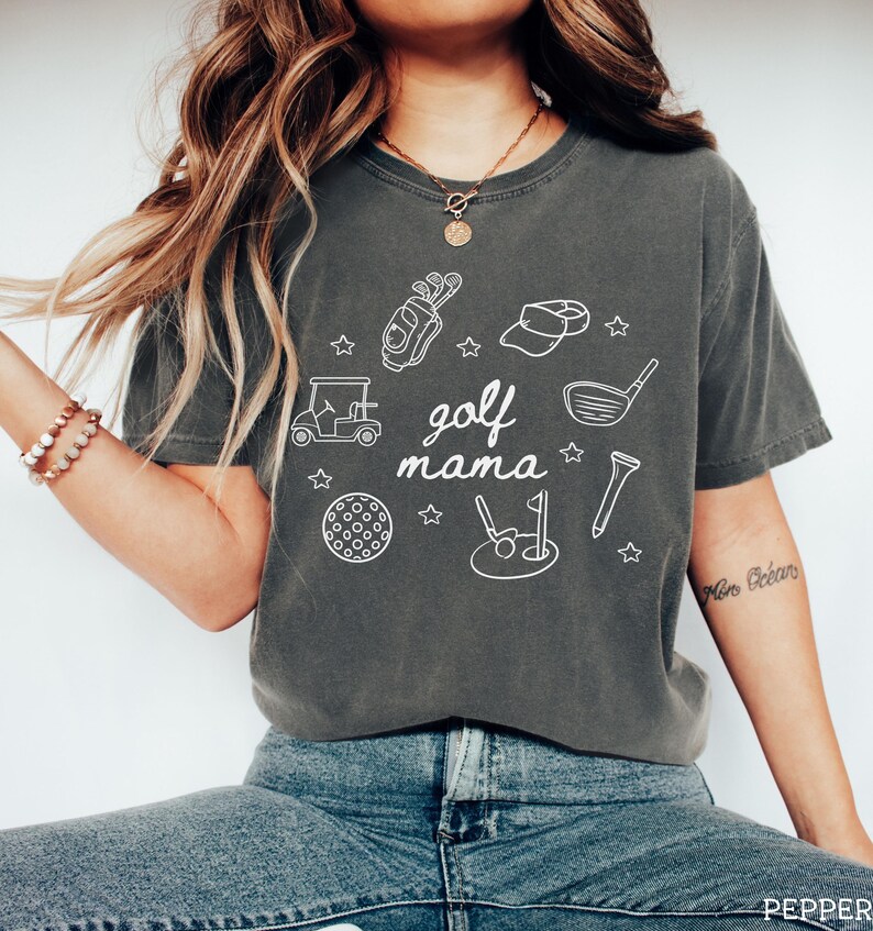 Puede incluir: Una camiseta gris con un dise&ntilde;o gr&aacute;fico blanco de art&iacute;culos relacionados con el golf, incluyendo un carrito de golf, palos de golf, una pelota de golf y una bandera de golf. El texto "golf mama" est&aacute; escrito en blanco en la camiseta.