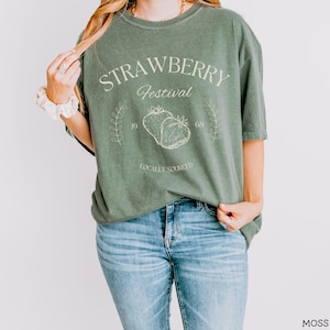 K&ouml;nnte beinhalten: Ein gr&uuml;nes T-Shirt mit dem Text "Strawberry Festival" und einer Grafik von zwei Erdbeeren. Der Text "Locally Sourced" befindet sich unter den Erdbeeren.