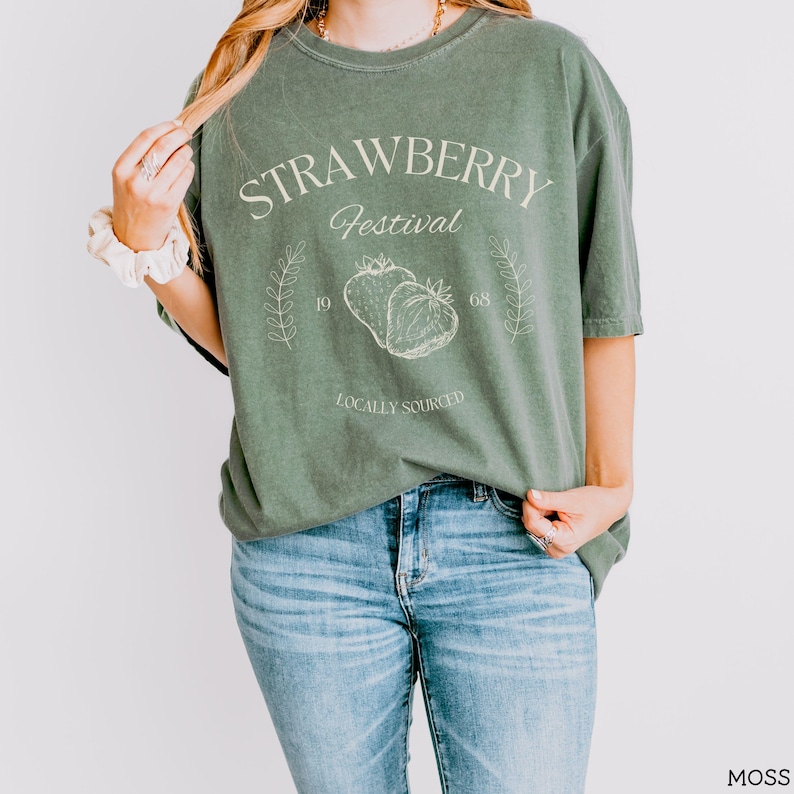 K&ouml;nnte beinhalten: Ein gr&uuml;nes T-Shirt mit dem Text "Strawberry Festival" und einer Grafik von zwei Erdbeeren. Der Text "Locally Sourced" befindet sich unter den Erdbeeren.
