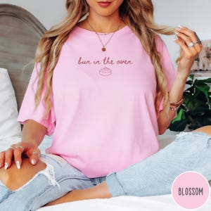 K&ouml;nnte beinhalten: Hellrosa T-Shirt mit dem Schriftzug "bun in the oven" in Schreibschrift und einer kleinen Zimtschnecken-Grafik. Das Shirt besteht aus einem weichen, bequemen Material.