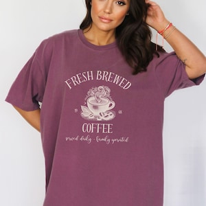 Op de afbeelding: Een pruimkleurig t-shirt met korte mouwen en een grafische print van een dampende kop koffie. De tekst luidt FRESH BREWED COFFEE, 1968 en served daily - family operated. Het ontwerp is in het midden op de borst in een lichte cr&egrave;mekleur.