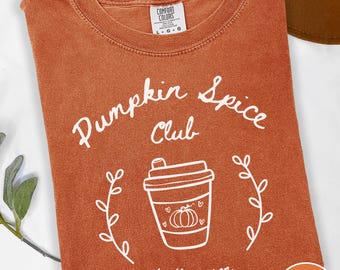 May include: A burnt orange short-sleeve t-shirt featuring white screen-printed text that reads Pumpkin Spice Club and tis the season. The design includes a line-art illustration of a coffee cup with a pumpkin and hearts, flanked by two leafy vines.