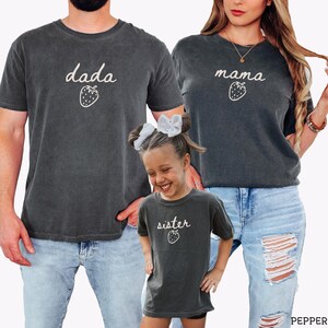 May include: Three charcoal gray short-sleeve t-shirts featuring white script text and a strawberry graphic. The shirts display the words "dada", "mama", and "sister" respectively, each accompanied by a small, simple line-drawn strawberry icon.