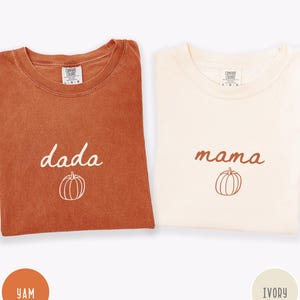 May include: Two Comfort Colors t-shirts, one in Yam and the other in Ivory. The Yam shirt has the word "dada" and a pumpkin graphic. The Ivory shirt has the word "mama" and a pumpkin graphic. Both shirts are folded.