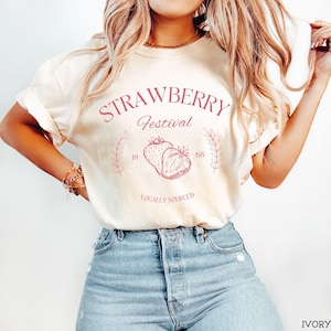 K&ouml;nnte beinhalten: Ein wei&szlig;es T-Shirt mit einem rosafarbenen Grafikdesign, auf dem "Strawberry Festival 19 68 Locally Sourced" steht. Die Grafik zeigt zwei Erdbeeren.