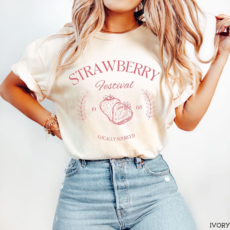 K&ouml;nnte beinhalten: Ein wei&szlig;es T-Shirt mit einem rosafarbenen Grafikdesign, auf dem "Strawberry Festival 19 68 Locally Sourced" steht. Die Grafik zeigt zwei Erdbeeren.