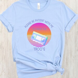 May include: A light blue t-shirt featuring a graphic of a VHS cassette tape centered over a sunset-colored circle. The text reads "PLEASE BE PATIENT WITH ME" above the graphic and "I'M FROM THE 1900'S" below the graphic in a purple sans-serif font.