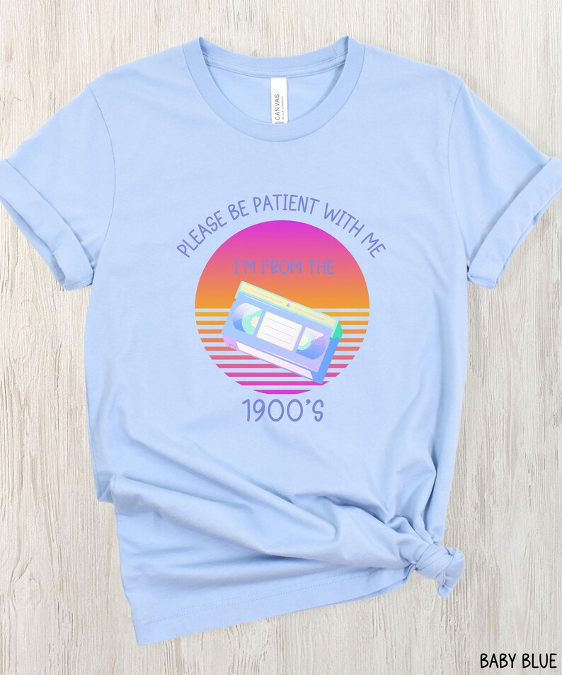 May include: A light blue t-shirt featuring a graphic of a VHS cassette tape centered over a sunset-colored circle. The text reads "PLEASE BE PATIENT WITH ME" above the graphic and "I'M FROM THE 1900'S" below the graphic in a purple sans-serif font.