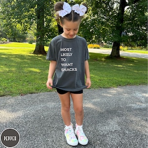 Puede incluir: Una camiseta gris carb&oacute;n de manga corta con el texto blanco MOST LIKELY TO WANT SNACKS impreso en el frente. La camiseta se combina con pantalones cortos negros y zapatillas altas con un patr&oacute;n tie-dye pastel multicolor.