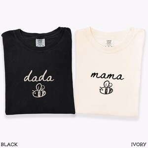 May include: Black and ivory t-shirts with the words "dada" and "mama" in cursive script, each with a bee graphic. The shirts are folded and displayed together. The words "BLACK" and "IVORY" are displayed below the shirts.