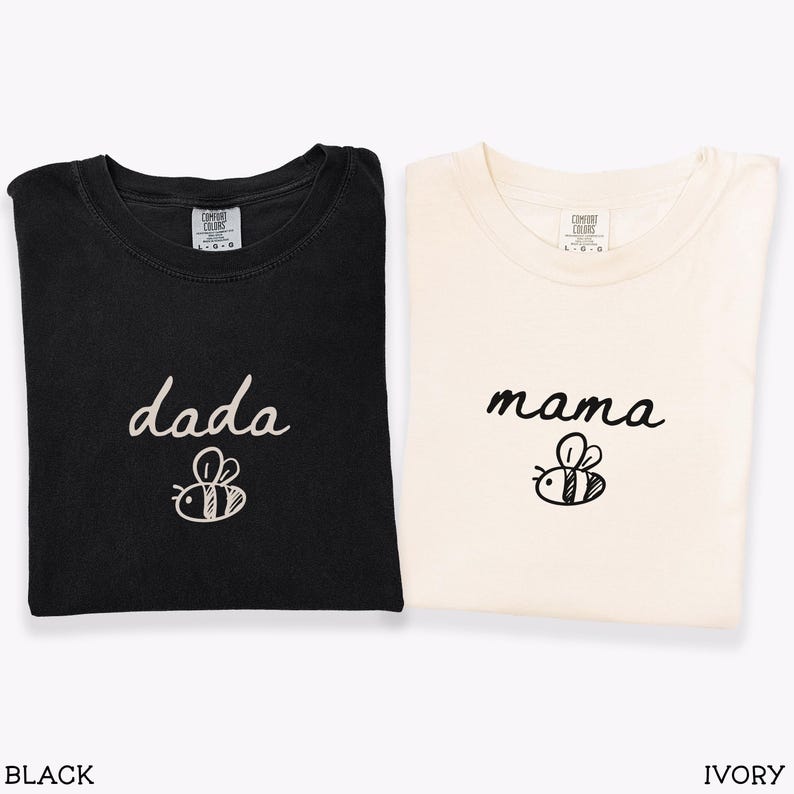 May include: Black and ivory t-shirts with the words "dada" and "mama" in cursive script, each with a bee graphic. The shirts are folded and displayed together. The words "BLACK" and "IVORY" are displayed below the shirts.