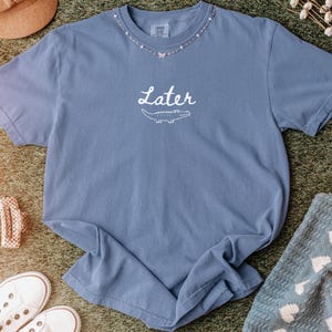 Pu&ograve; includere: Una maglietta blu jeans con la scritta "Later" e una grafica di un alligatore. La maglietta ha uno scollo tondo con un design a collana di perline. La maglietta &egrave; realizzata in un materiale morbido ed &egrave; appoggiata su una superficie verde.