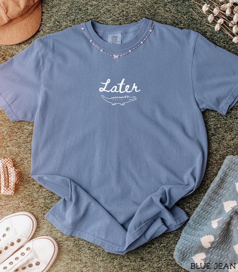 Pu&ograve; includere: Una maglietta blu jeans con la scritta "Later" e una grafica di un alligatore. La maglietta ha uno scollo tondo con un design a collana di perline. La maglietta &egrave; realizzata in un materiale morbido ed &egrave; appoggiata su una superficie verde.