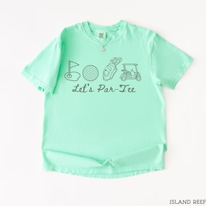 May include: A mint green t-shirt with a graphic design featuring golf-related illustrations and the text "Let's Par-Tee". The design includes a golf flag, golf ball, golf bag, and golf cart. The shirt is made of a soft material.