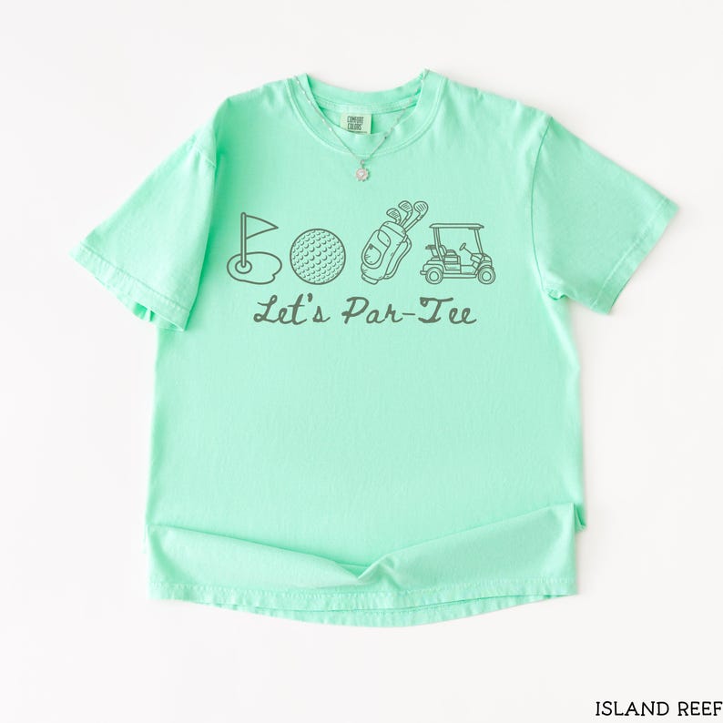 May include: A mint green t-shirt with a graphic design featuring golf-related illustrations and the text "Let's Par-Tee". The design includes a golf flag, golf ball, golf bag, and golf cart. The shirt is made of a soft material.